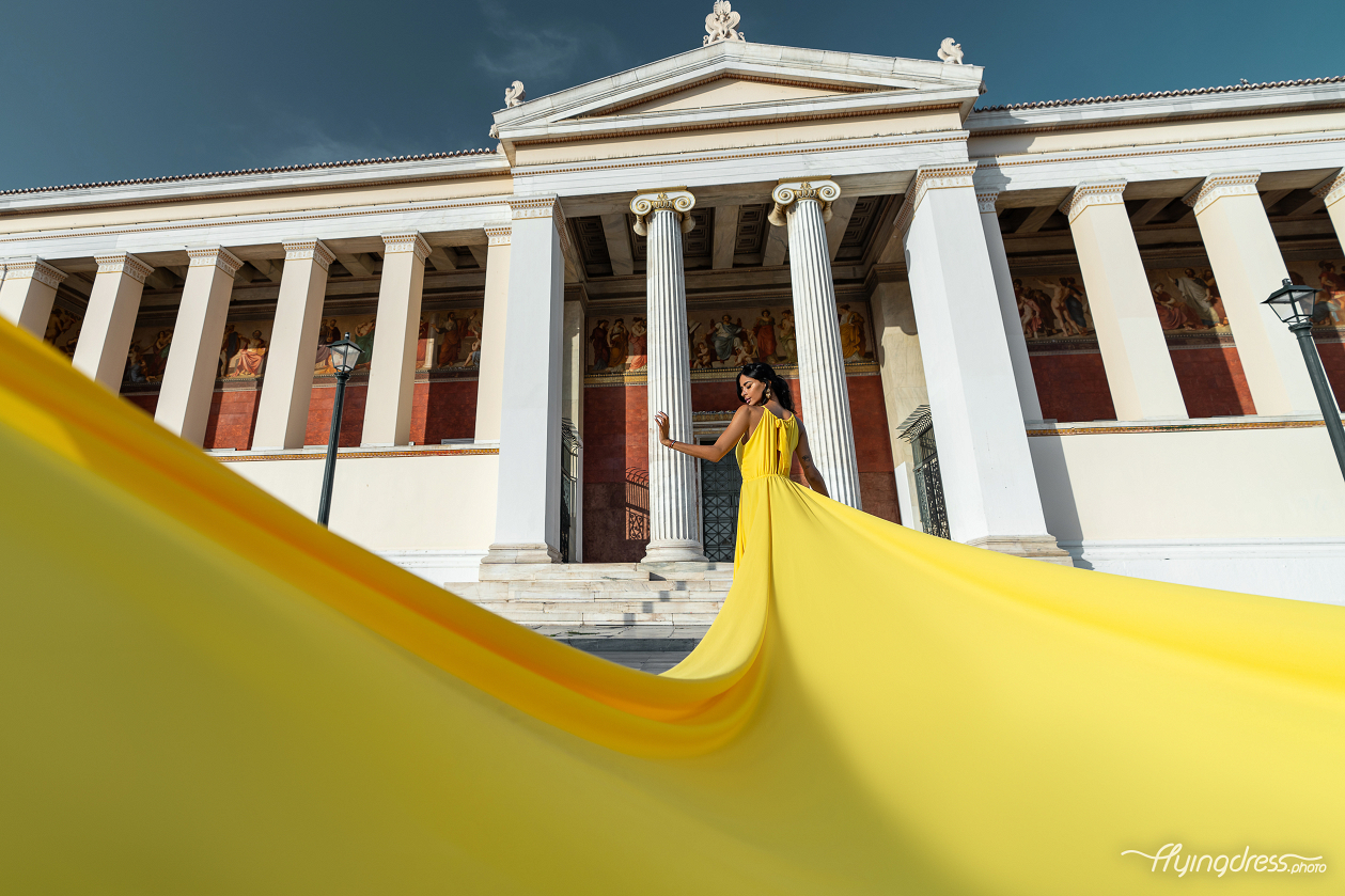 A woman in a yellow dress stands gracefully on the steps of Athens University, with her dress flowing dramatically behind her, framed by the building’s grand columns and vibrant murals.